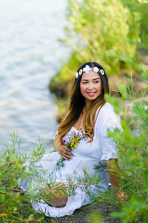 pregnant woman wearing flower crown and a white maternity dress for her maternity shoot near the river